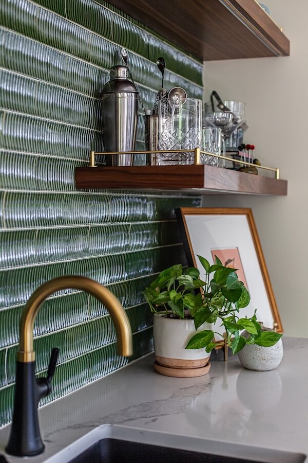 A kitchen countertop with a potted plant, framed art, and a gold faucet below a shelf holding glassware and bar tools against a green tiled backsplash reflects upcoming interior design trends 2026.