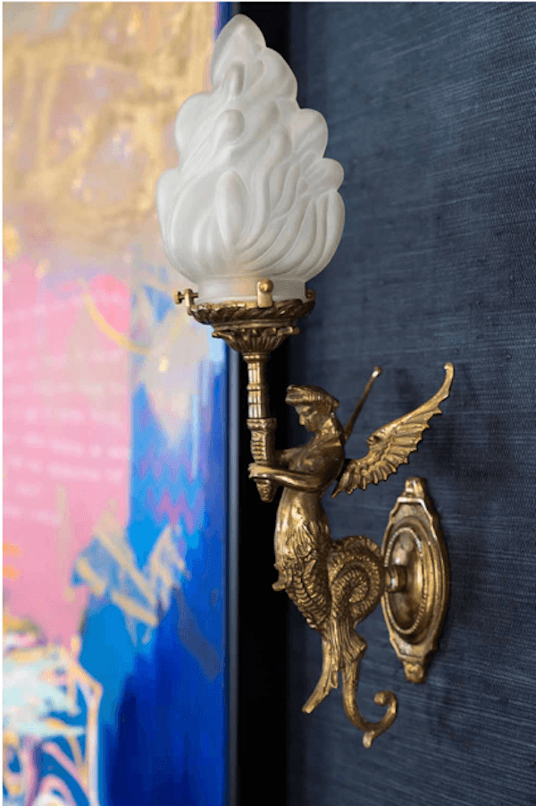 A brass wall sconce shaped like a winged figure holding a torch, with a frosted flame-shaped glass shade, adds a touch of anticipated interior design trends 2026 when mounted on a dark wall.