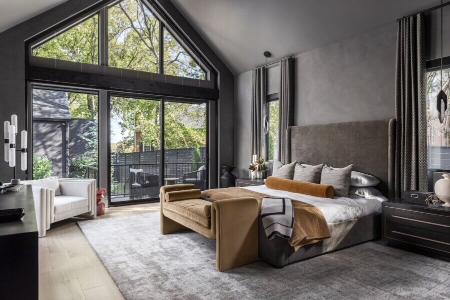 A modern bedroom with large floor-to-ceiling windows, a neutral color scheme, an upholstered bed, a tan bench, and a view of trees outside—perfectly reflecting interior design trends 2026.