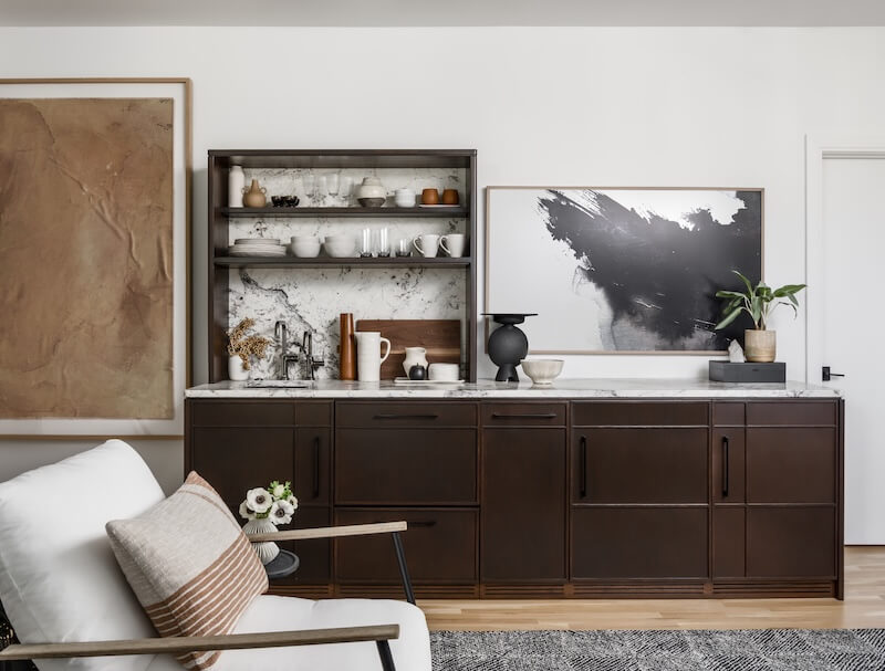 A modern kitchen features dark wood cabinets, open shelving with dishes, a marble backsplash, abstract art, and decorative items next to a white armchair with neutral pillows—showcasing interior design trends 2026.