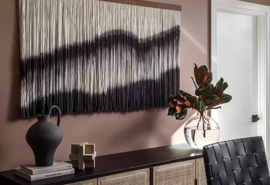 Wall art made of hanging yarn in gradient black and white, a nod to interior design trends 2026, displayed above a sideboard with a black vase, decorative object, and a glass vase holding leafy branches.