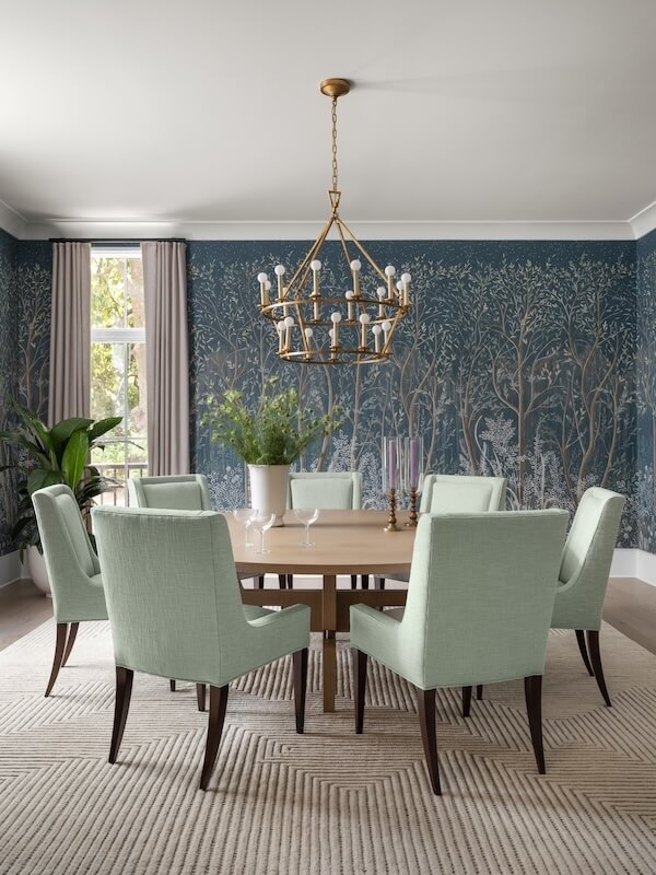 A dining room inspired by 2026 interior design trends features a wooden table, eight light green upholstered chairs, a gold chandelier, and blue tree-patterned wallpaper. Green plants and elegant curtains complete this stylish interior design.