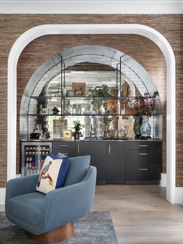 A modern home bar with black cabinetry, open glass shelves, and decorative items showcases 2026 interior design trends. Set within an arched alcove, a blue swivel chair with a bird pillow adds charm to this stylish home decor scene.