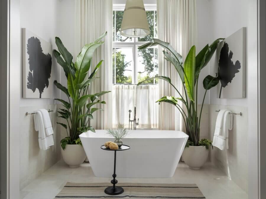 Embracing 2026 Interior Design trends, this modern bathroom features a freestanding white bathtub, lush potted green plants, two towels, a round side table, white curtains, and a window welcoming natural light.