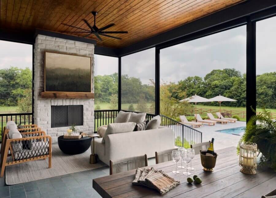 A covered patio with a seating area, fireplace, and dining table overlooks a pool with lounge chairs and umbrellas, surrounded by greenery—reflecting interior design trends 2026 that blend indoor comfort with outdoor elegance.