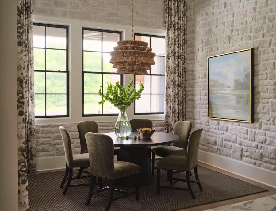 A small dining area with a round black table, five upholstered chairs, a large glass vase with green branches, and windows with patterned curtains reflects interior design trends 2026, set against light brick walls.
