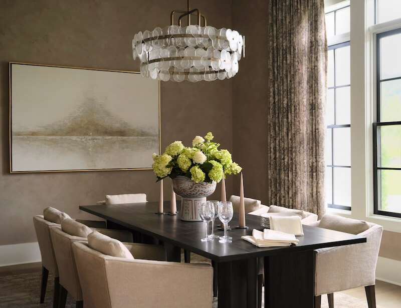 A modern dining room featuring a dark rectangular table, eight beige chairs, a large floral centerpiece, a glass chandelier, and a neutral abstract painting highlights interior design trends 2026 with its elegant style and large windows with drapes.