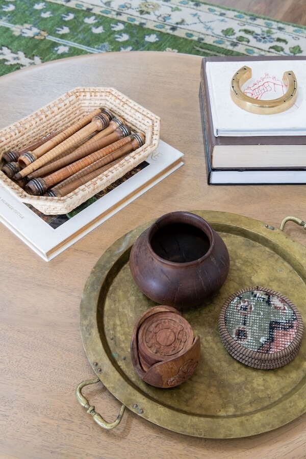 A wooden table with a brass tray holding a carved wooden container and a round embroidered box; nearby are stacked books, a woven basket with spools, and part of a green rug, reflecting interior design trends 2026.