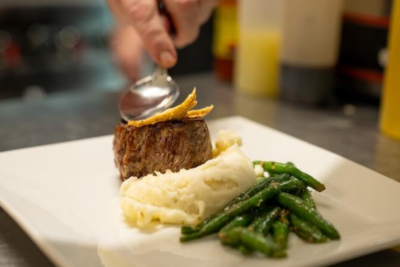 A hand uses a spoon to garnish a plated meal of steak, mashed potatoes, and green beans on a white square dish—perfect fuel before exploring top Mississippi destinations.