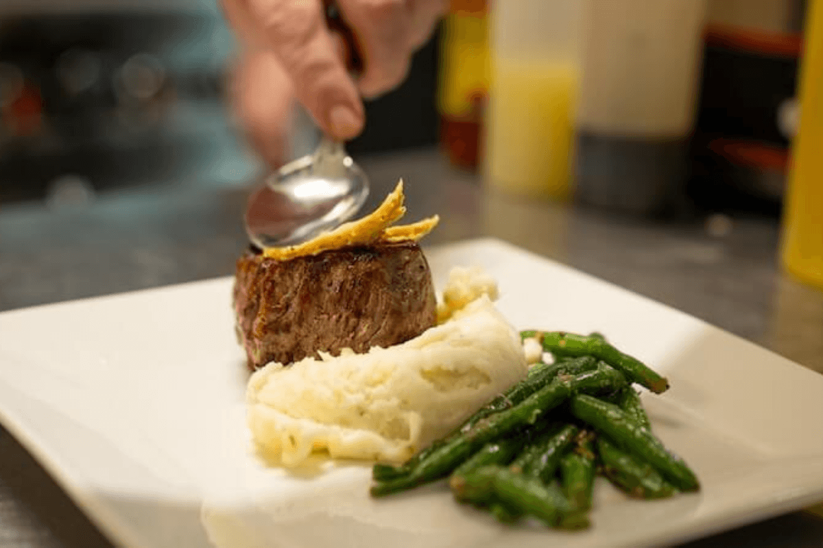 A hand uses a spoon to garnish a plated meal of steak, mashed potatoes, and green beans on a white square dish—perfect fuel before exploring top Mississippi destinations.