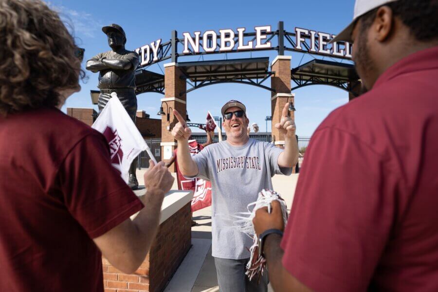 8 Mississippi Destinations Worth the Drive - 7 Three people wearing Mississippi State gear stand outside Dudy Noble Field, a favorite spot among Mississippi destinations. One person in the center smiles and raises their hands, with a statue and stadium entrance sign visible behind them.