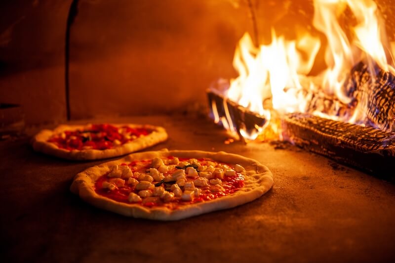 8 Mississippi Destinations Worth the Drive - 16 Two uncooked pizzas with cheese and tomato sauce are baking inside a wood-fired oven with flames visible on the right side, capturing the rustic charm found in many Mississippi destinations.