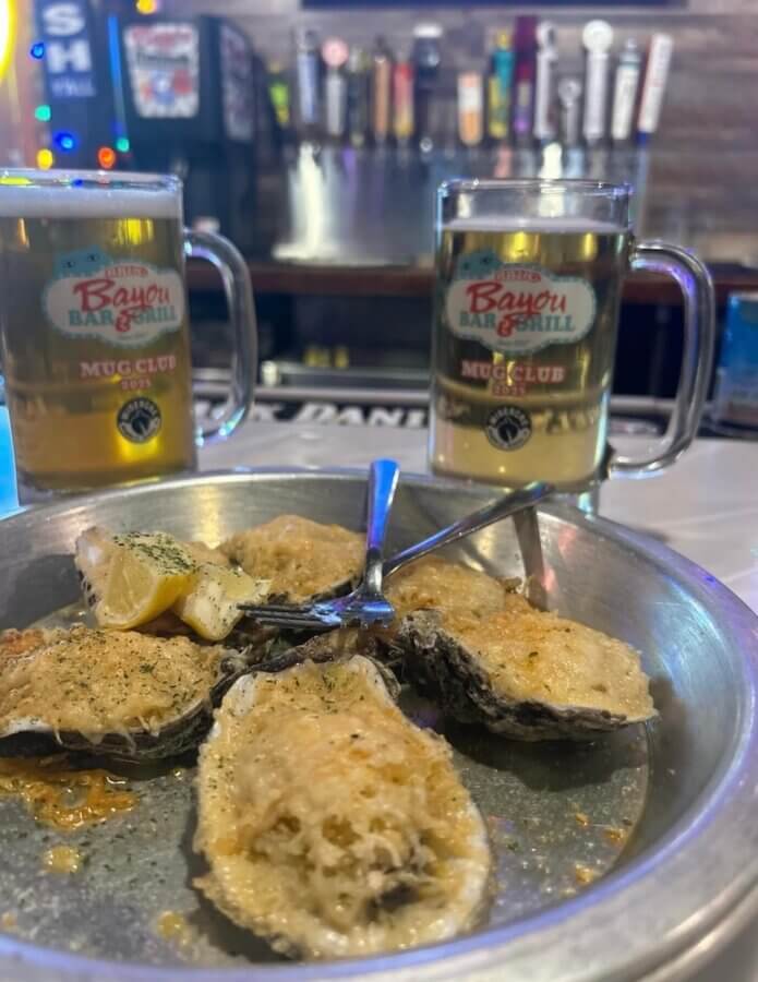 13 Spots to Grab Dinner at the Bar in Memphis - 12 A metal plate with baked oysters and lemon wedges sits in front of two beers in Bayou Bar & Grill mugs on a bar counter, capturing the vibrant atmosphere of one of the best bars in Memphis.