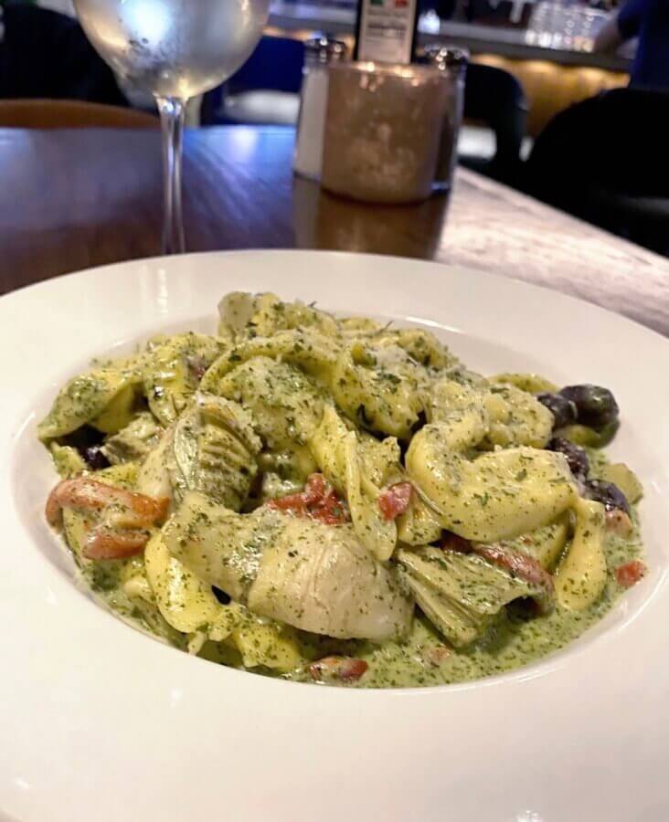 13 Spots to Grab Dinner at the Bar in Memphis - 4 A white bowl filled with pasta in a creamy green sauce with artichokes and vegetables, perfect for dinner at a Memphis bar, sits beside a glass of white wine and salt and pepper shakers in the background.