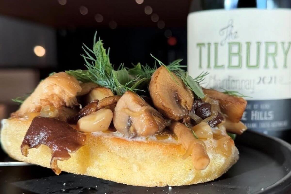 A slice of bread topped with cooked mushrooms and fresh herbs sits on a black plate next to a bottle labeled "The Tilbury," making it one of the best spots for dinner at the bar in Memphis.