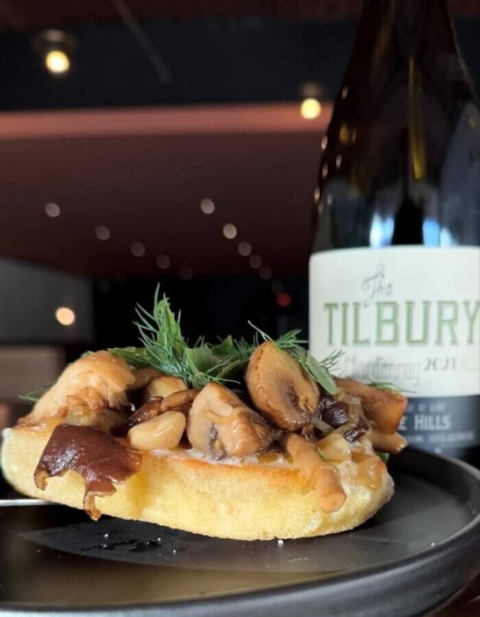 13 Spots to Grab Dinner at the Bar in Memphis - 10 A slice of toasted bread topped with cooked mushrooms and herbs sits on a plate, capturing the essence of Memphis dining and hinting at one of the best spots for dinner at the bar, with "The Tilbury Chardonnay 2021" in the background.