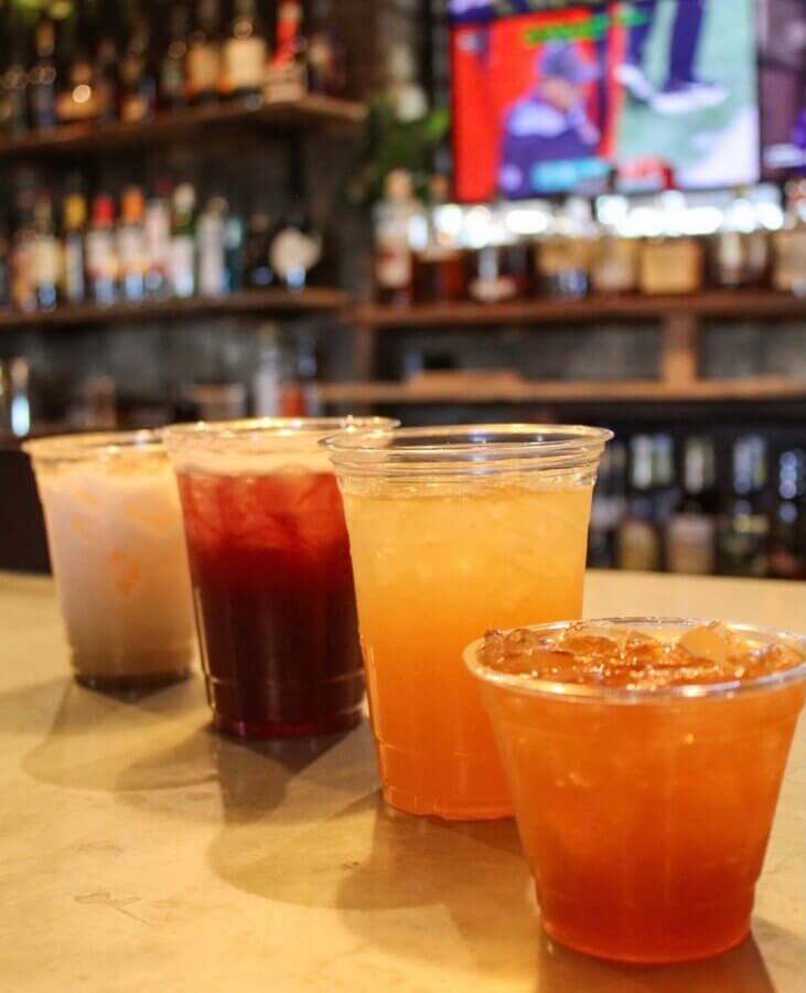 13 Spots to Grab Dinner at the Bar in Memphis - 3 Four different cocktails in plastic cups are arranged on a bar counter at a lively Memphis bar, with shelves of bottles and a TV in the background.