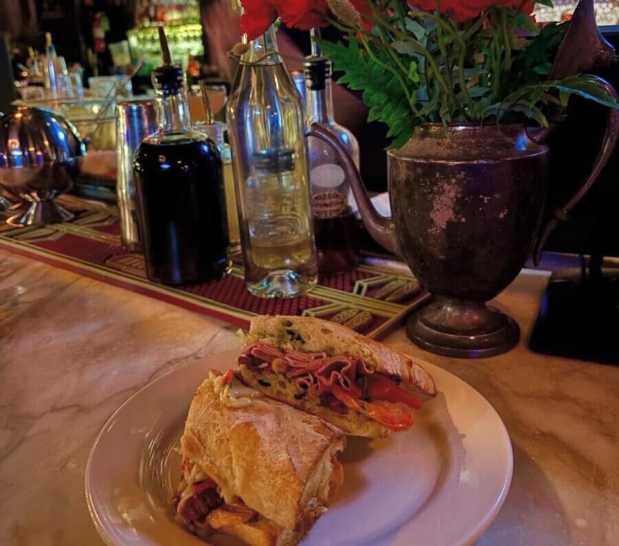 13 Spots to Grab Dinner at the Bar in Memphis - 5 A sandwich on a white plate sits on a marble bar counter in Memphis, with bottles of oil and vinegar and a metal vase with red flowers in the backgroundβone of the best spots for dinner at the bar.