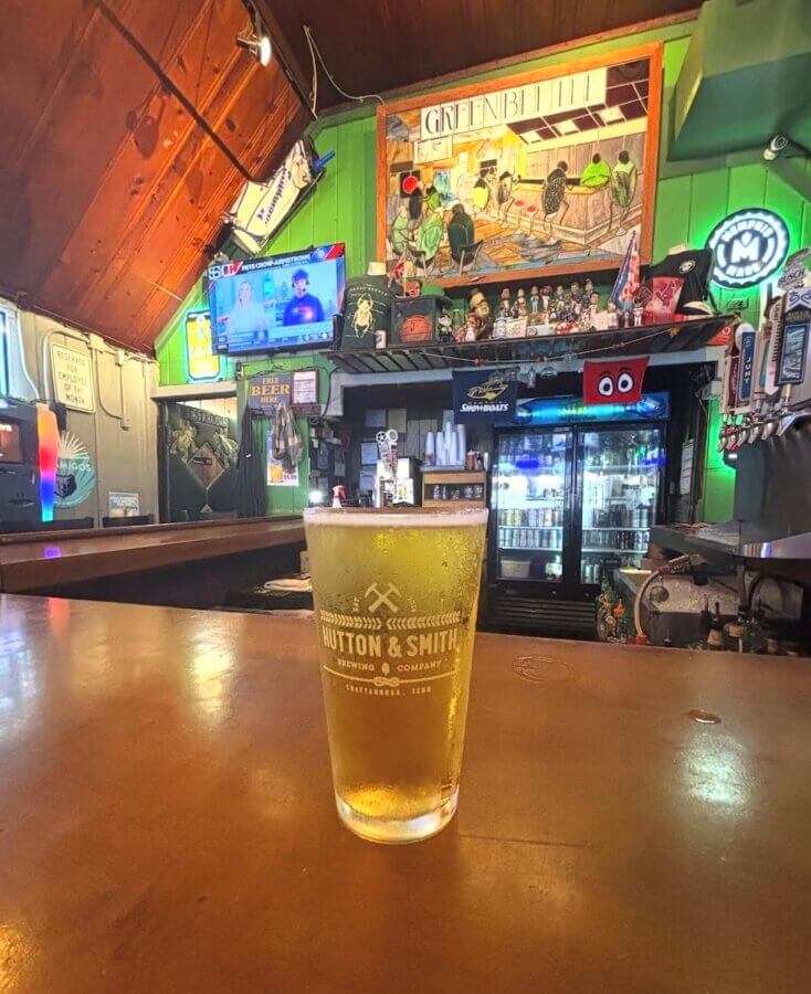 13 Spots to Grab Dinner at the Bar in Memphis - 8 A pint of beer in a Hutton & Smith glass sits on a wooden bar inside a rustic Memphis pub, with a TV, fridge, and colorful wall art in the backgroundβperfect for relaxing before dinner.