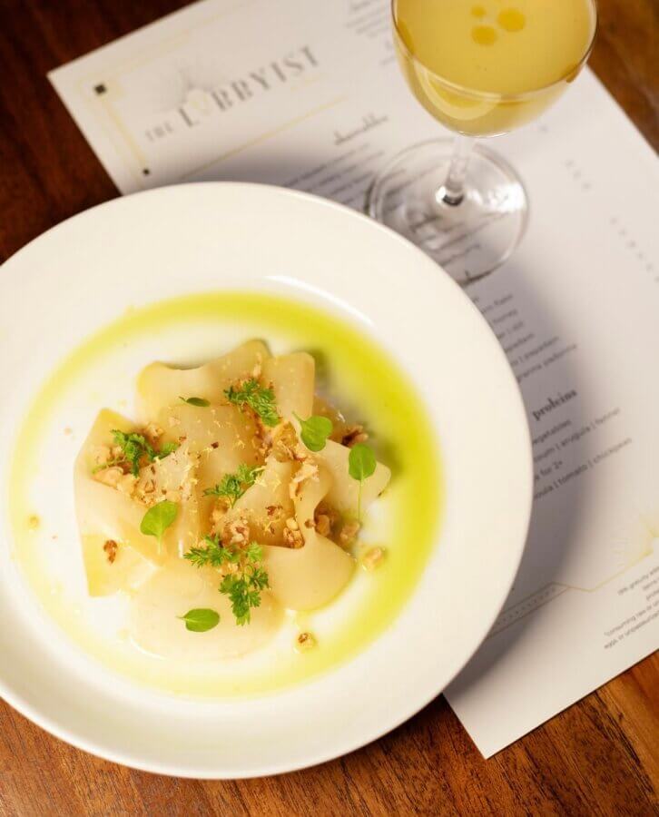 13 Spots to Grab Dinner at the Bar in Memphis - 6 A white plate with thinly sliced scallops, garnished with herbs and nuts, sits next to a yellow cocktail on a bar menu atop a wooden tableβperfect for a stylish Memphis dinner.