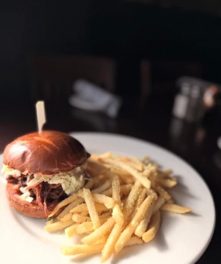 13 Spots to Grab Dinner at the Bar in Memphis - 11 A plate with a Memphis-style pulled pork sandwich topped with coleslaw on a brioche bun and a serving of French fries, perfect for dinner at your favorite bar.