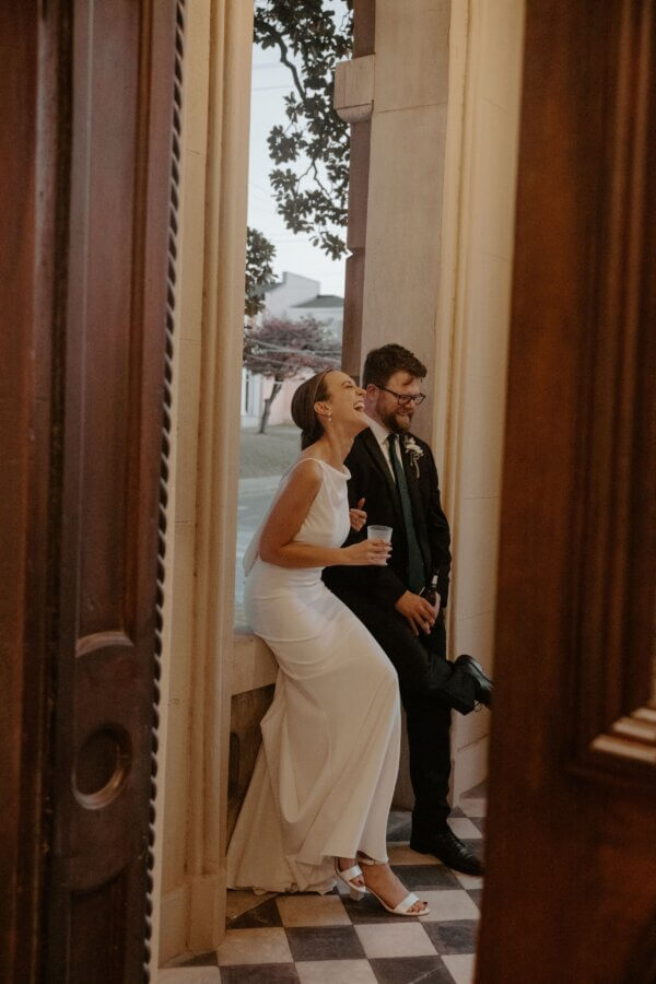An Elegant Memphis Wedding at The Historic James Lee House - 19 A bride in a white dress and a groom in a black suit sit laughing together by a window, holding drinks, at the Historic James Lee House with checkered floors.