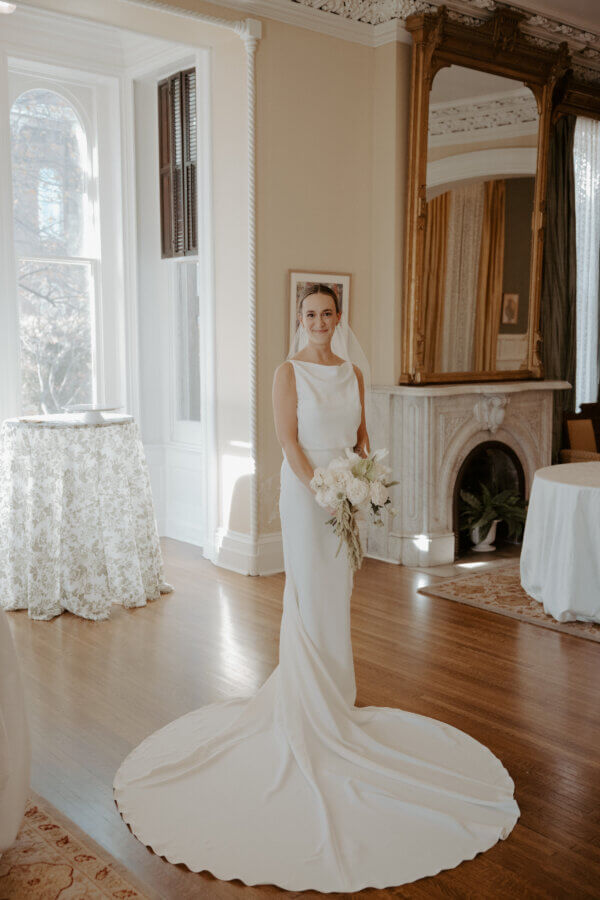 An Elegant Memphis Wedding at The Historic James Lee House - 6 A bride stands in a stately bridal suite wearing a simple gown and white bouquet.