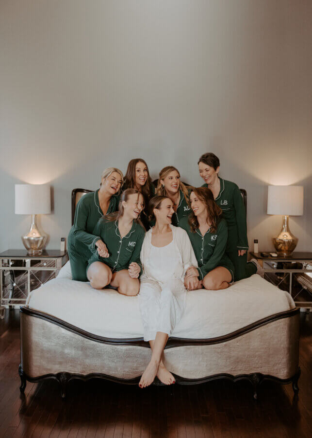 An Elegant Memphis Wedding at The Historic James Lee House - 9 A bride wearing white pajamas sits in the middle of her bridesmaids wearing green pajamas on a bed smiling for a group photo.