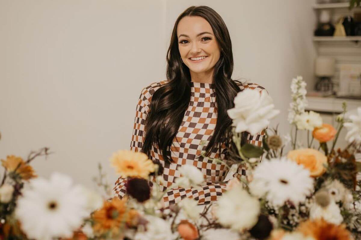 A woman with long dark hair, possibly Kallie Holstein, wearing a checkered dress sits at a table decorated with assorted flowers in a bright indoor setting, her face illuminated by natural light.