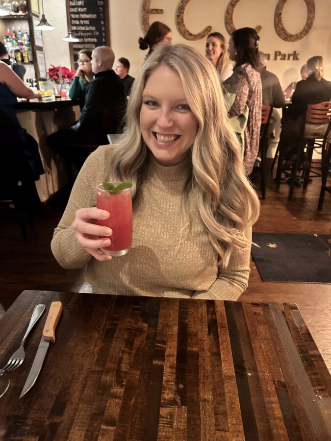 Meet Jasmin Day of Ease Wellness & JDay Bakes - 2 A blond woman sits at a restaurant table, holding a drink.