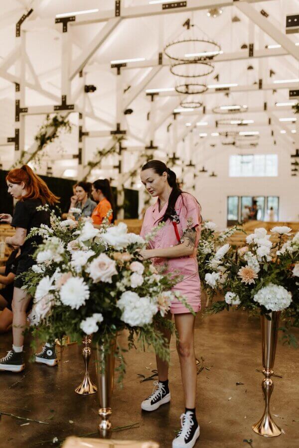 Meet Kallie Holstein of Dainty Florals - 4 Kallie Holstein, in a pink dress, arranges flowers in a bright, spacious venue with white beams and floral decorations.
