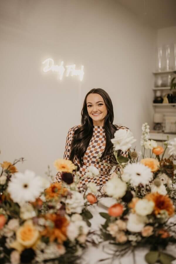 Meet Kallie Holstein of Dainty Florals - 3 A woman with long dark hair sits behind a table decorated with colorful floral arrangements in a well-lit room with a neon sign on the wall.
