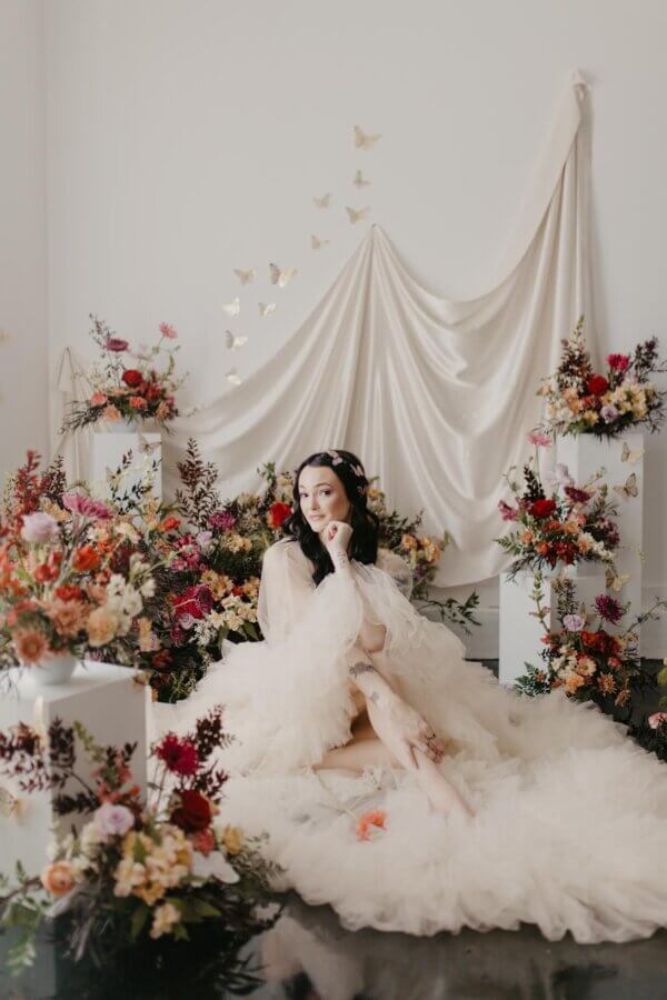 Meet Kallie Holstein of Dainty Florals - 2 A woman in a fluffy white dress sits on the floor surrounded by colorful floral arrangements and draped fabric with butterfly decorations on a white wall behind her.