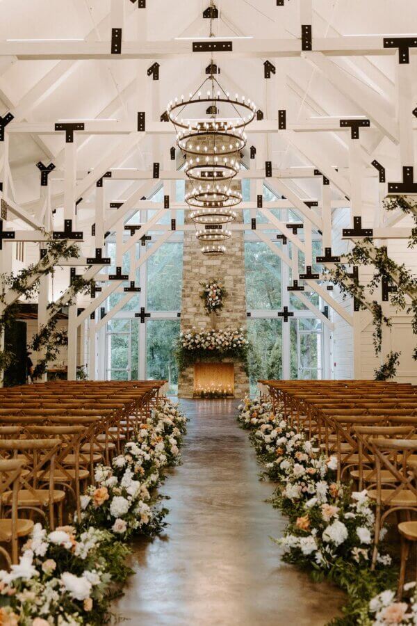 Meet Kallie Holstein of Dainty Florals - 5 A wedding ceremony space features wooden chairs, floral aisle decorations, chandeliers, and a stone fireplace adorned with flowers at the altar.