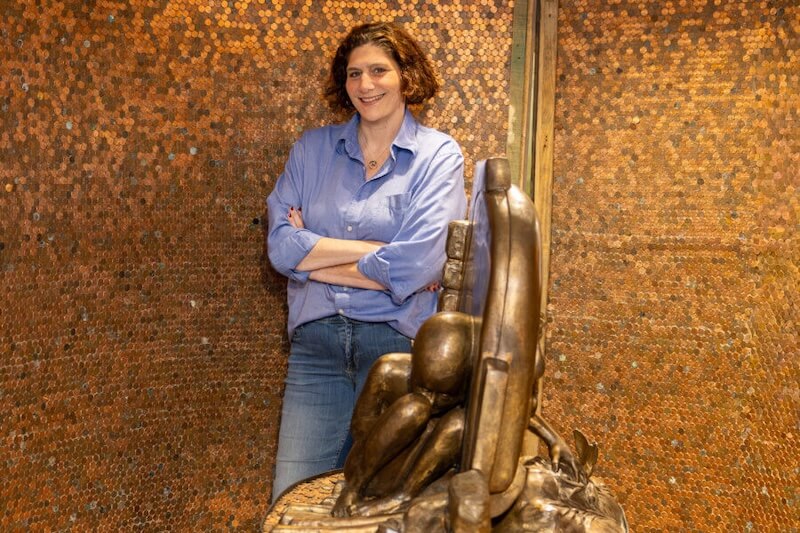A woman with curly hair stands with arms crossed in front of a bronze sculpture of an unknown child, with a wall covered in pennies behind her.