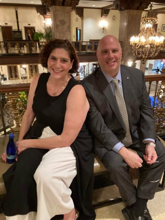 Honoring the 1.5 Million: Leah Fox-Greenberg and the Mission of the Unknown Child Foundation - 5 Leah Fox-Greenberg and a man in a suit sit side by side on a bench in an elegant, chandelier-lit lobby, their FACES illuminated by the soft glow.