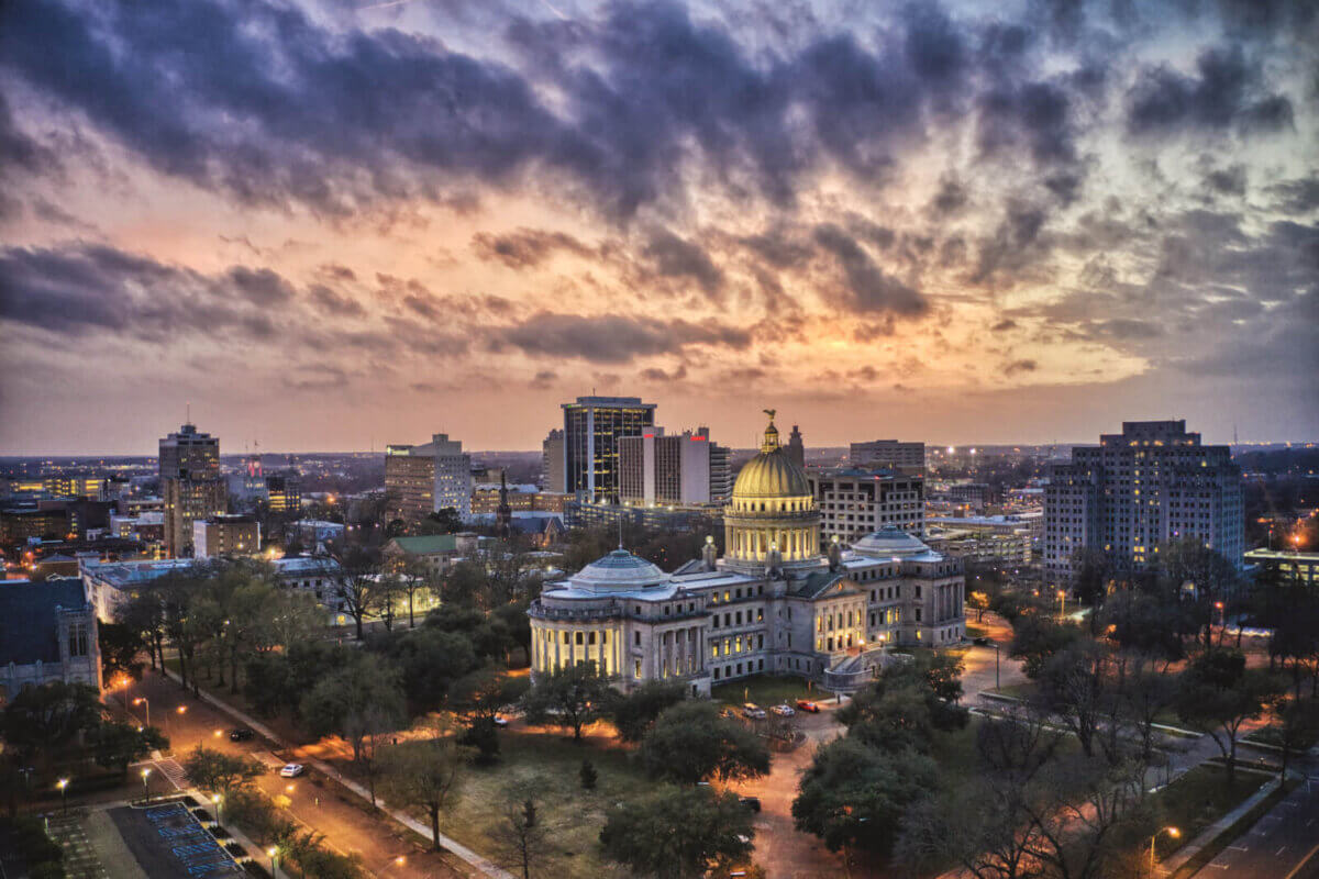 Aerial view of the Mississippi State Capitol building surrounded by city buildings in Jackson at sunset, with dramatic clouds in the sky, highlighting nearby Jackson neighborhoods and renowned Jackson restaurants.