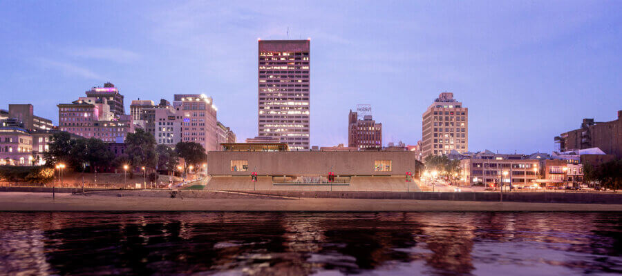 New to Memphis! Exciting Additions for 2026 - 2 A modern, rectangular building sits on the Memphis riverbank with a skyline of mid-rise city buildings in the background at dusk—an exciting preview of what's coming soon in 2026.