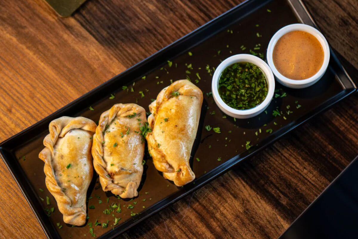 Three baked empanadas on a rectangular black plate with two small bowls of dipping sauces, garnished with chopped herbs—a delicious choice at one of the top new Memphis restaurants.