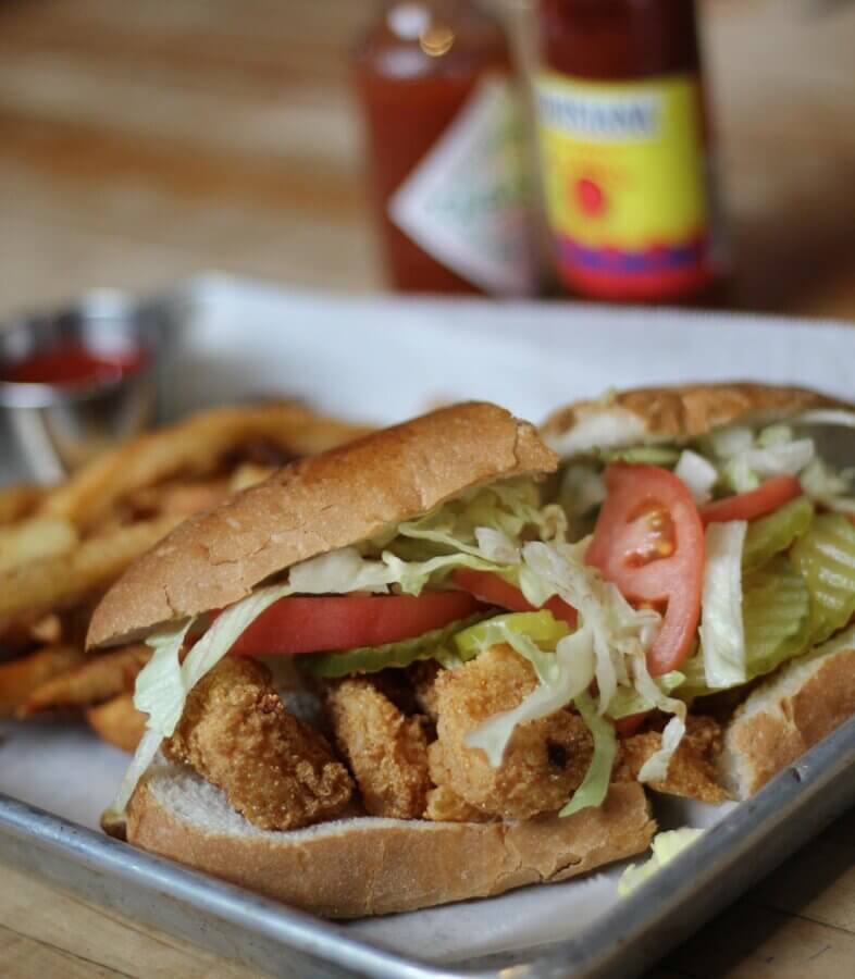 New to Memphis! Exciting Additions for 2026 - 6 A fried shrimp po' boy sandwich with lettuce, tomato, and pickles is served on a tray with French fries and condiments in the background. Coming soon to Memphis in 2026!.