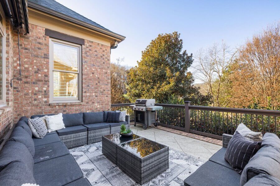 Just Listed! A Franklin Home Designed to Entertain & Unwind - 11 Outdoor patio of this Franklin home features gray sectional seating, decorative pillows, a glass-top coffee table, rug, and gas grill—perfectly designed to entertain—surrounded by trees and a charming brick exterior.