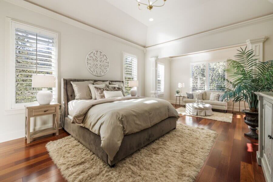 Just Listed! A Franklin Home Designed to Entertain & Unwind - 12 Bright bedroom in a Franklin Home with a large bed, beige bedding, two rugs, wooden floors, and a comfy seating area—perfect to entertain or unwind. Enjoy natural light from multiple windows with shutters and a lush potted plant. Just listed!.