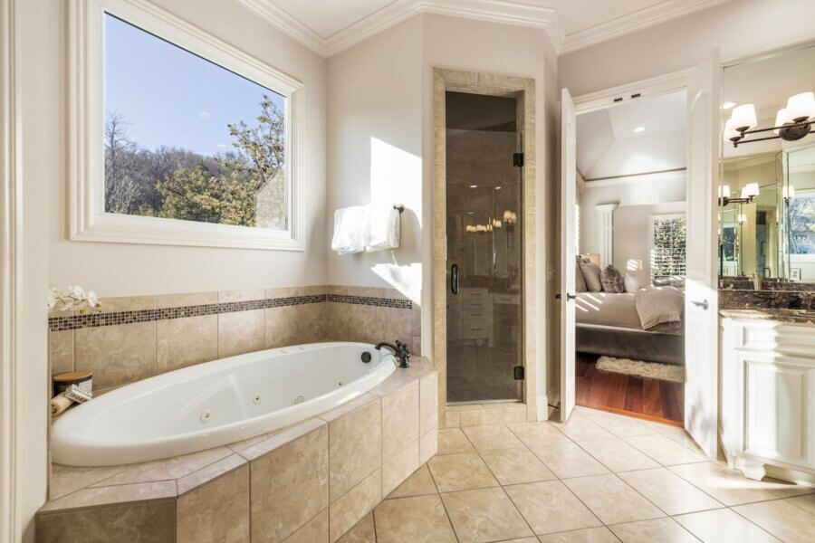 Just Listed! A Franklin Home Designed to Entertain & Unwind - 13 Modern bathroom in a Franklin Home with beige tile flooring, a large built-in bathtub, glass shower, and double doors leading to the bedroom; sunlight streams through a window. Just listed and perfect for relaxing or entertaining guests.