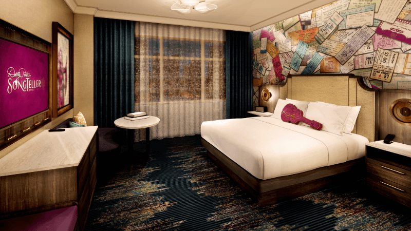 Hotel room in Nashville with a king bed, guitar-shaped pillow, TV displaying “Songteller,” small table and two chairs, dark curtains, and a wall decorated with colorful 2026 concert ticket stubs.