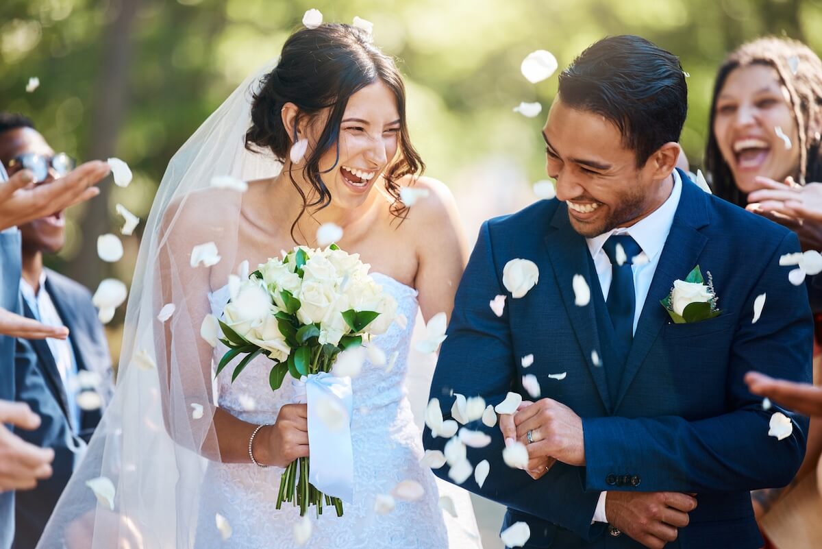 A bride and groom, both smiling, walk outside surrounded by people throwing white flower petals. The bride holds a bouquet of white roses and wears a white dress and veil—perfect inspiration for any Nashville wedding resource.