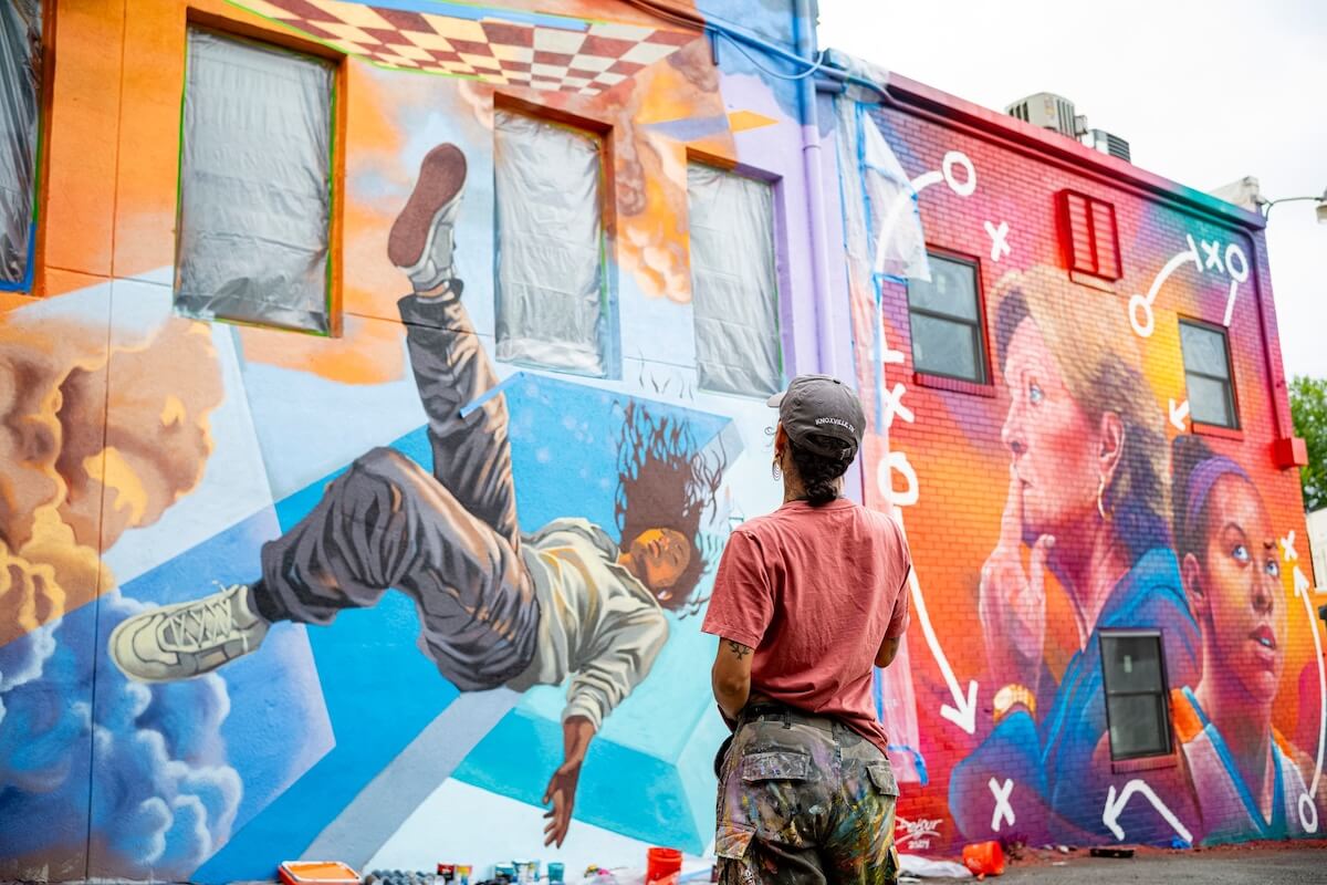 An artist stands in front of a colorful mural featuring a falling figure and two faces with white symbols drawn over the vibrant background, capturing the spirit of exciting developments in the south.