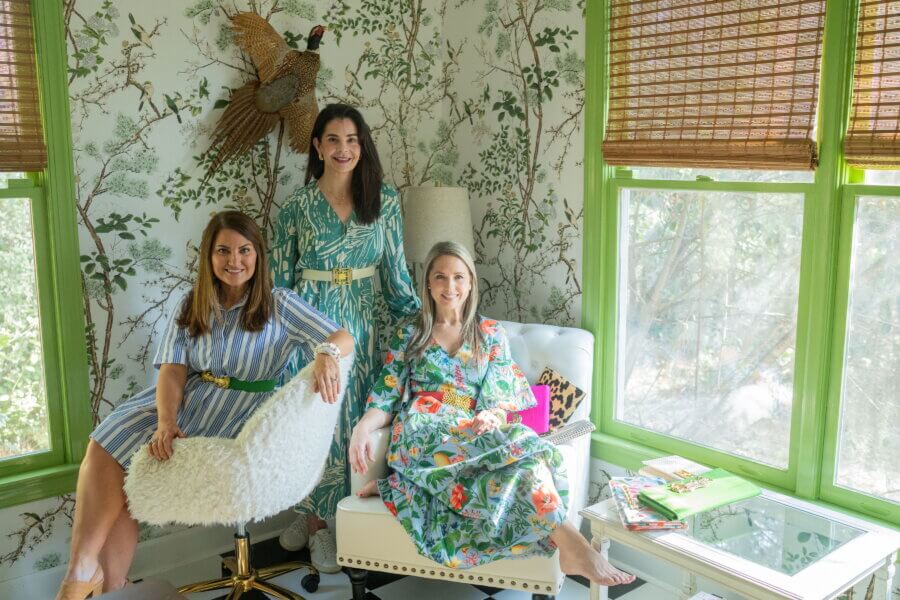 Meet Lindsay Thomas: Founder of Garland Bags - 4 Three women sit and stand in a bright, green-trimmed room with tree-patterned wallpaper, wearing colorful dresses. Books and a lamp are on the table beside them.
