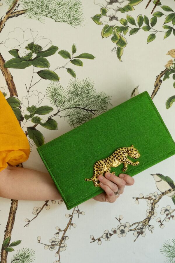 Meet Lindsay Thomas: Founder of Garland Bags - 2 A person in a yellow sleeve holds a green Garland Bags clutch with a gold leopard detail in front of floral-patterned wallpaper.
