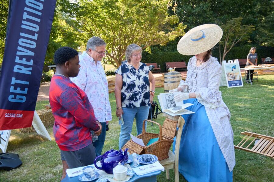 Coming in 2026! Exciting Developments to Watch Around the South - 10 A woman in historical costume shows papers to three people at an outdoor event with a “Get Revolution” banner and a table set with dishes and a teapot, discussing exciting developments for 2026.