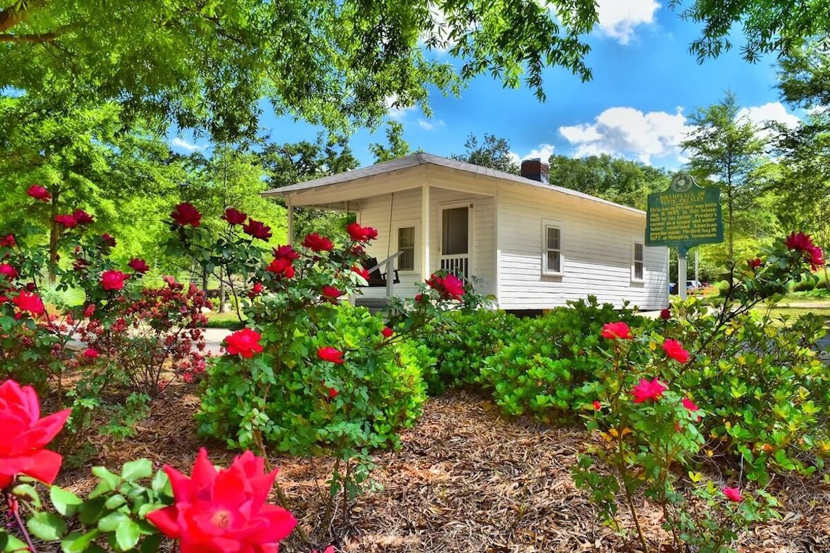 A small white house with a porch sits behind blooming red rose bushes and green trees on a sunny Mississippi day. A historical marker sign is visible on the right, inviting visitors to explore during Spring Break.